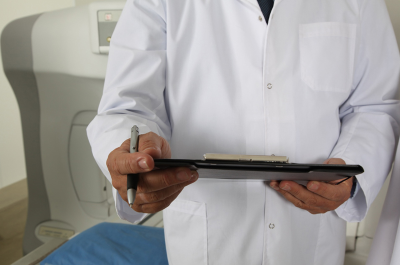 A shot of a doctor in a white coat holding a clipboard and a pen. His face is cut off from the frame.